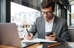 Smiling business man in eyeglasses sitting by the table in cafe with laptop computer while using smartphone and writing something