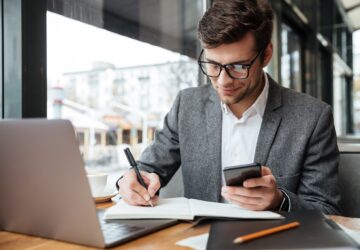 Smiling business man in eyeglasses sitting by the table in cafe with laptop computer while using smartphone and writing something